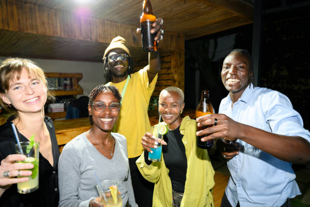 Waist-up view of casually dressed men and women in 20s and 30s holding beer and cocktails, laughing, smiling, and toasting camera. in a bar in Nairobi